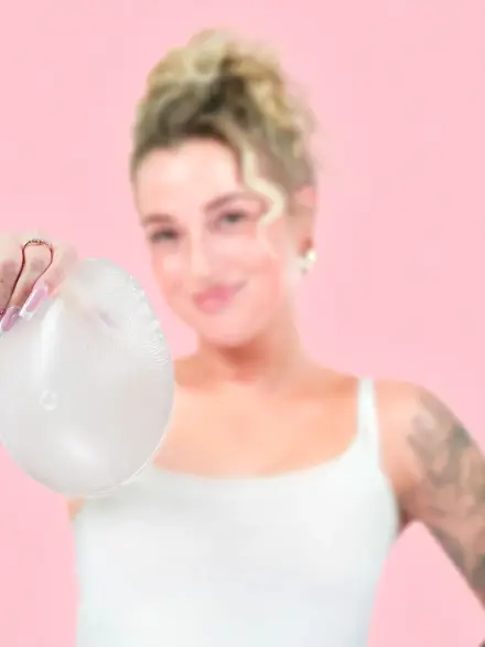 Frau in weißem Top hält eine transparente Silikonprothese in die Kamera; rosa Hintergrund, unscharfer Porträt-Look.